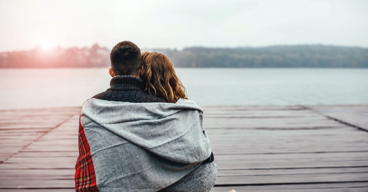 A couple wrapped in a blanket on a riverside dock at sunset
